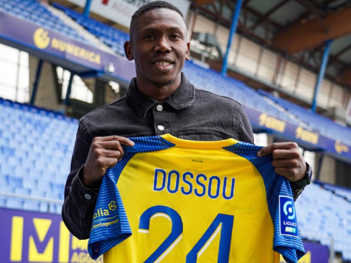 Un homme souriant tient un maillot de football jaune et bleu avec le nom "DOSSOU" et le numéro 24 dans un stade vide