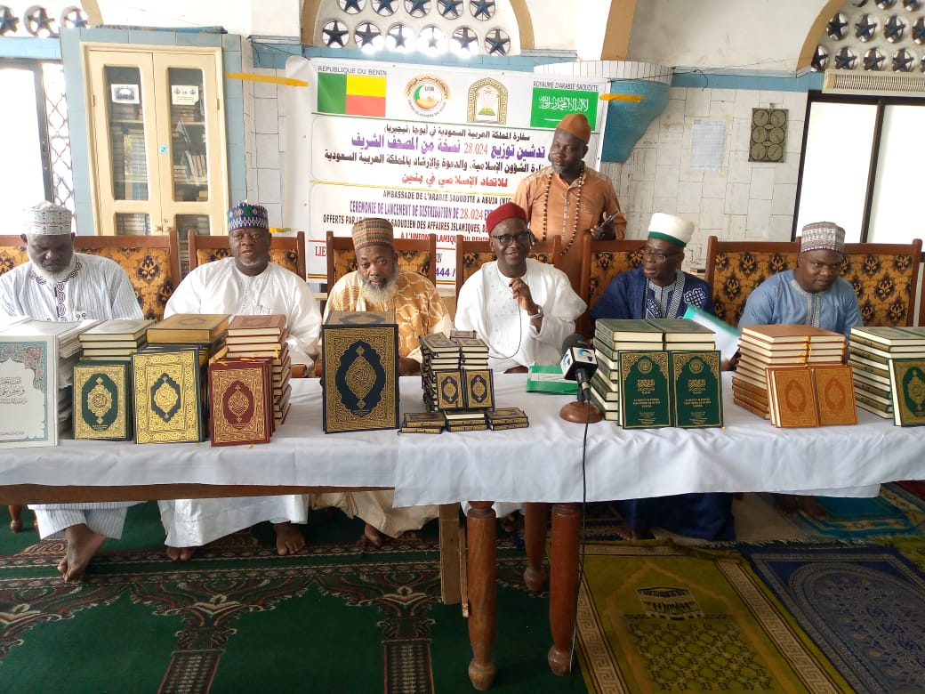 Plusieurs hommes en tenue traditionnelle sont assis derrière une table couverte de nombreux exemplaires du Saint Coran devant une bannière affichée au mur