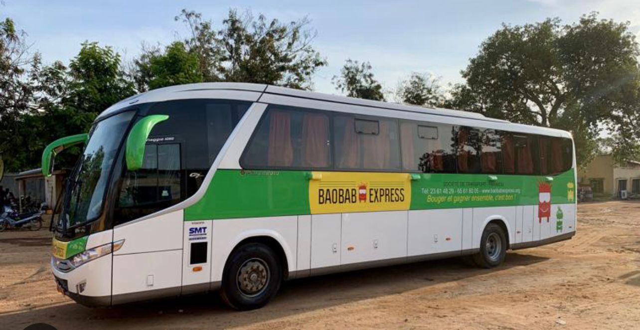 Bus blanc et vert avec la marque Baobab Express stationné sur un sol en terre entouré d’arbres et sous un ciel clair