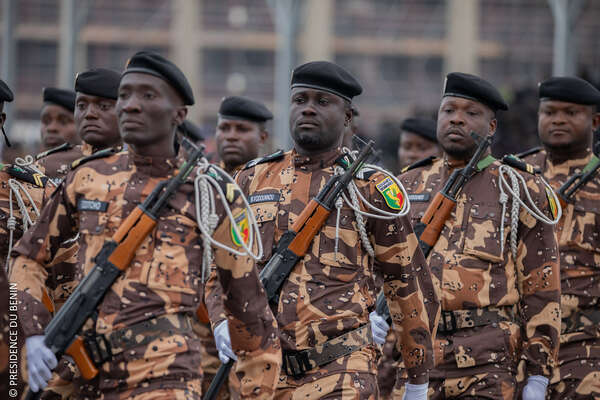 Des hommes en uniforme militaire à motifs camouflés avec bérets noirs et fusils en formation dans une rue urbaine.