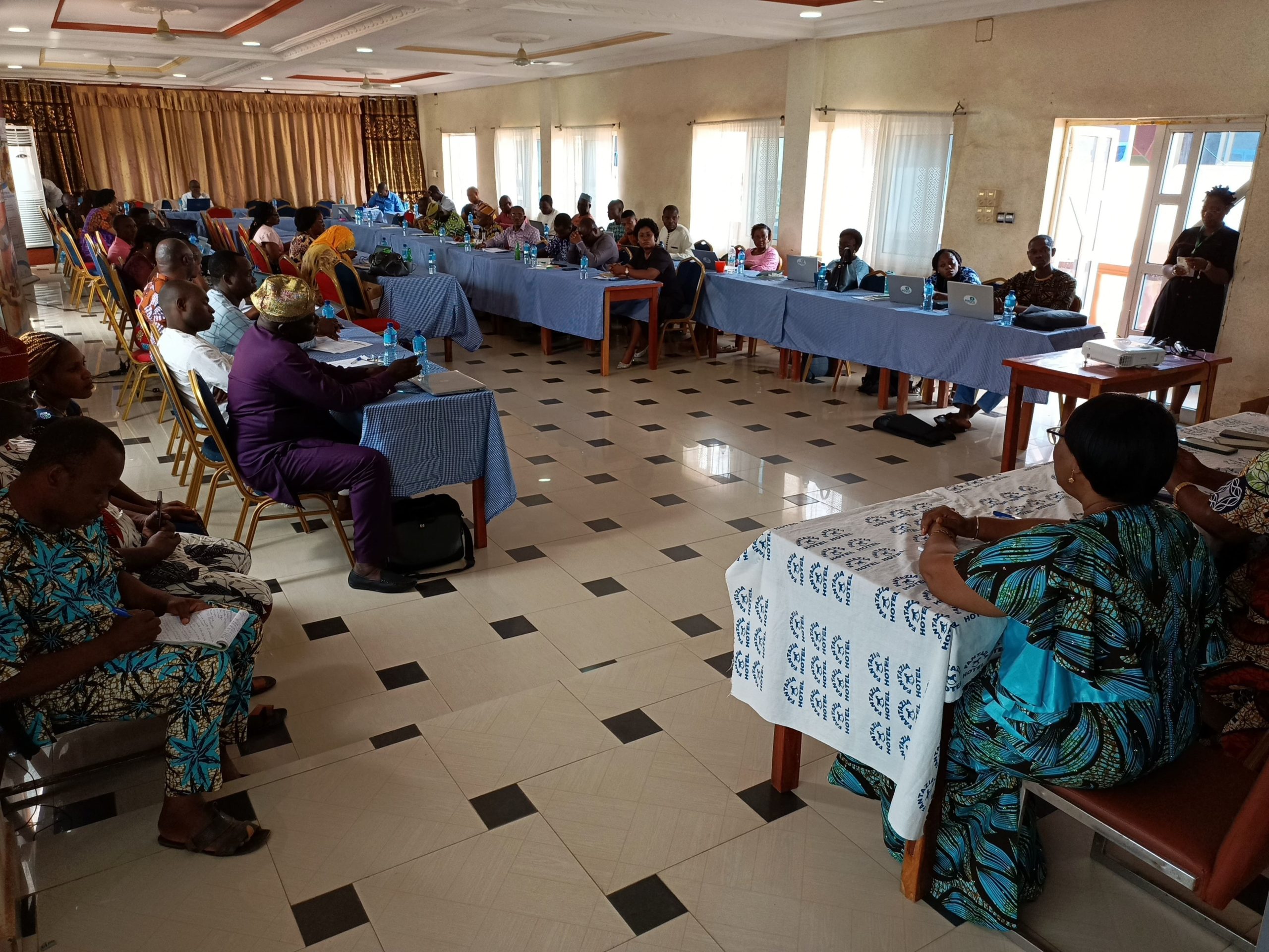 Un groupe de personnes assises en demi-cercle participent à une réunion dans une salle lumineuse avec des nappes bleues et blanches sur les tables