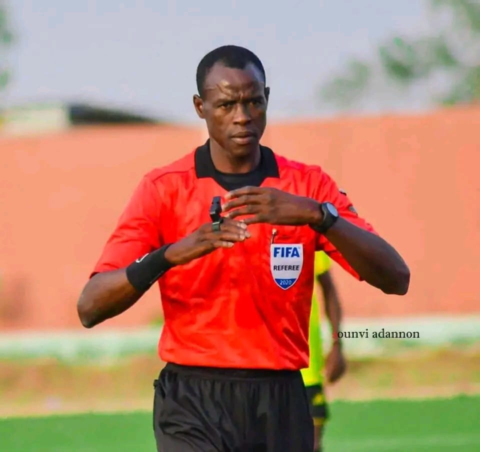 Arbitre en tenue rouge et noire ajustant un sifflet lors d’un match sur un terrain de football vert