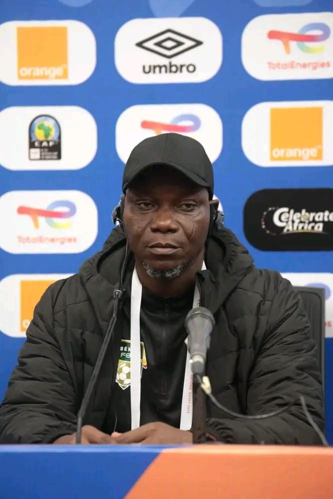 Homme assis devant un micro avec casquette noire et veste matelassée lors d'une conférence de presse sportive