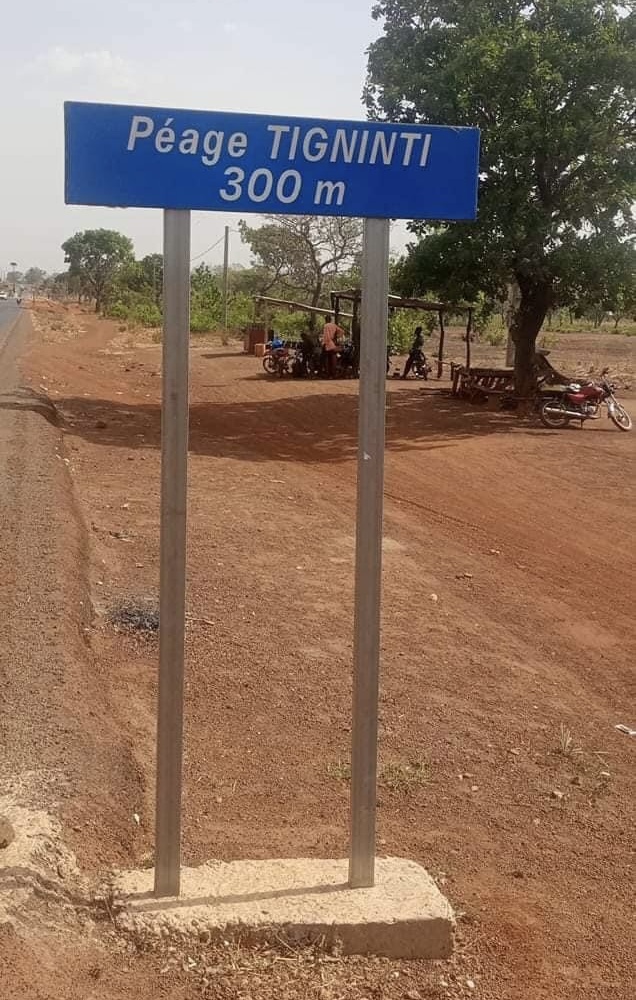 Panneau indiquant un péage à 300 mètres sur une route bordée de terre et quelques motos garées sous un arbre