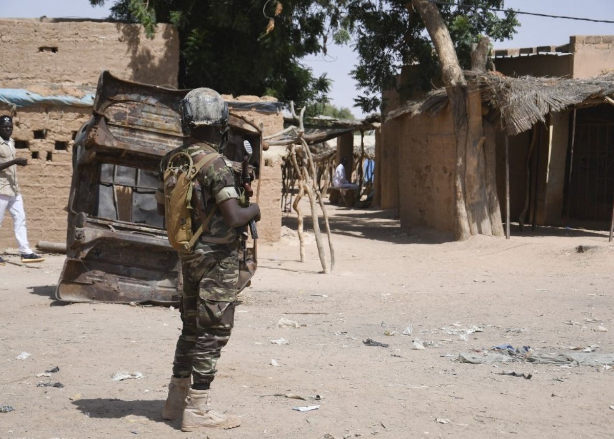 Militaire en tenue camouflée et casque tenant une arme dans un village désert avec bâtiments en terre et arbres autour