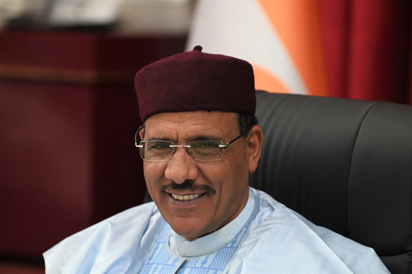 Homme souriant portant une tenue traditionnelle claire et un chapeau bordeaux assis sur un fauteuil noir
