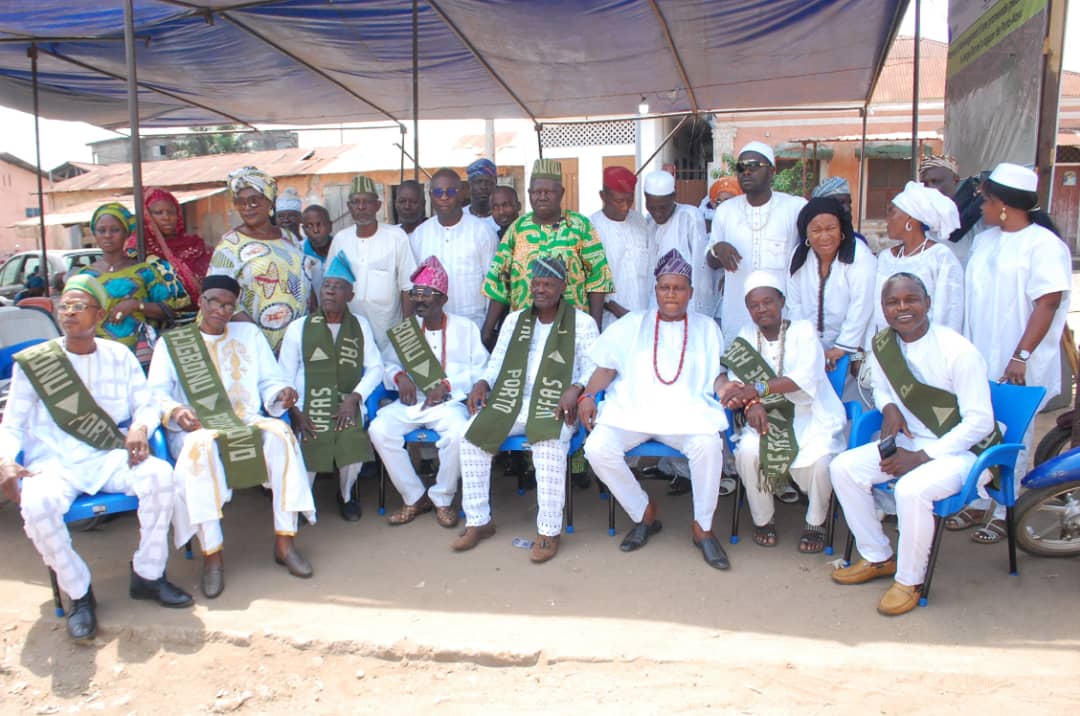 Groupe d’hommes et femmes posant sous un abri, certains assis avec des écharpes vertes portant des inscriptions blanches, vêtements traditionnels et modernes mê