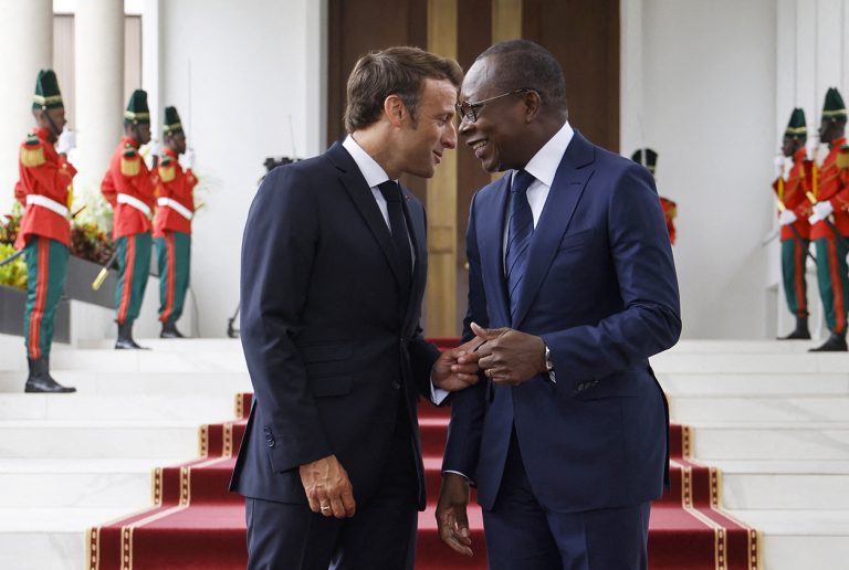 Deux hommes en costume échangent en souriant devant un escalier rouge orné de gardes en uniforme vert et rouge.