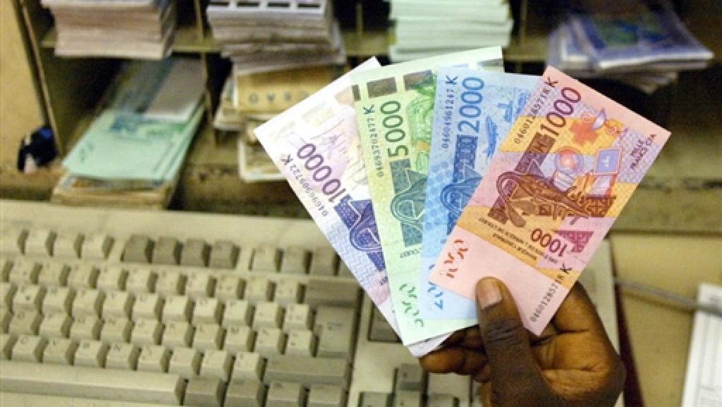 Main tenant plusieurs billets de banque colorés devant un clavier d’ordinateur ancien posé sur un bureau.