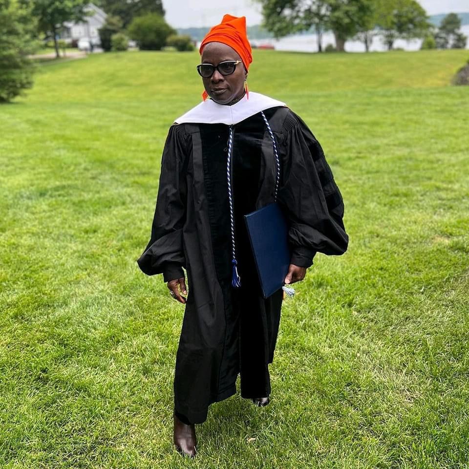Personne en toge noire et coiffe orange marchant sur une pelouse verte tenant un diplôme bleu