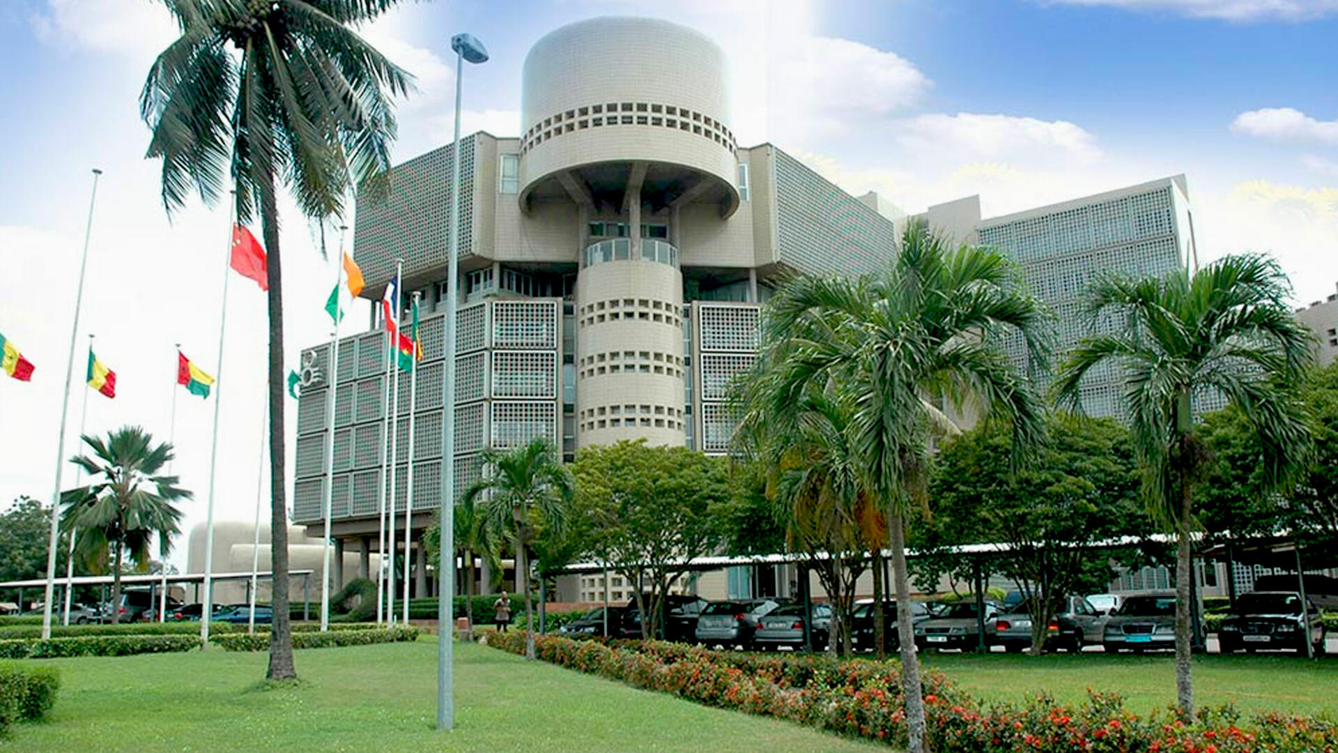 Bâtiment administratif moderne entouré de pelouses, arbres tropicaux et plusieurs drapeaux sur des mâts devant l’entrée