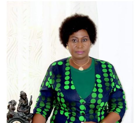 Une femme portant une robe à motifs verts et bleus posant devant un mur clair avec une statuette décorative noire à côté