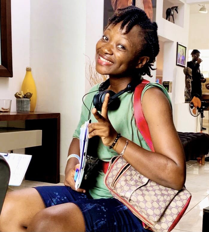 Jeune femme souriante en tenue décontractée assise avec un sac à main et un casque audio autour du cou faisant un geste de paix