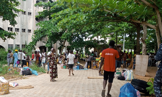Personnes rassemblées sous des arbres avec des sacs et affaires éparpillés sur un chemin en pavés