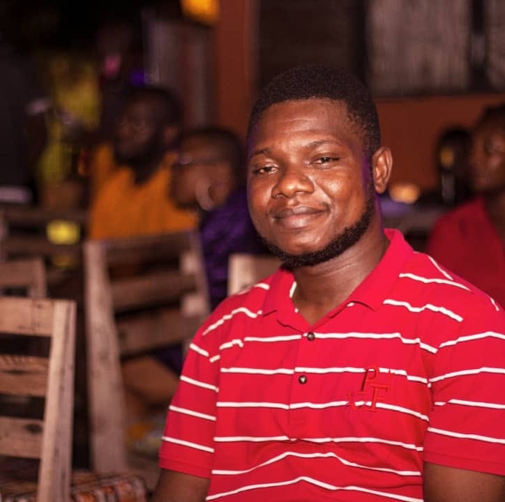 Homme souriant portant un polo rouge à rayures blanches assis dans un lieu intérieur avec des chaises en bois autour