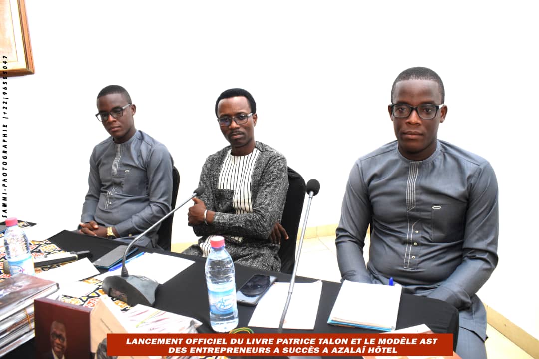 Trois hommes assis côte à côte derrière une table avec des micros et des documents face à un mur blanc
