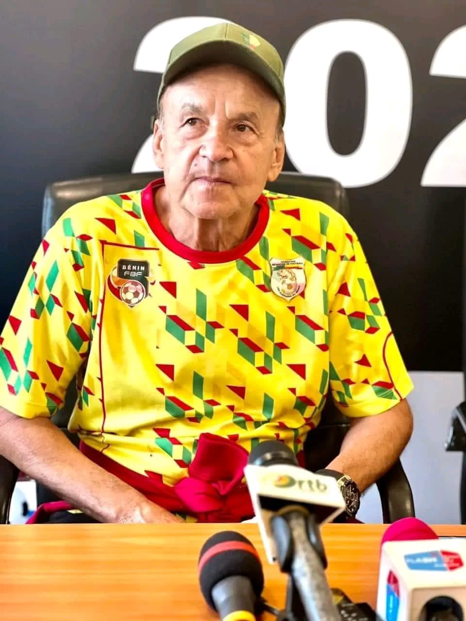 Homme âgé en tenue colorée avec logo sportif assis devant table avec micros lors d’une conférence