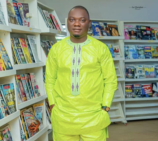 Homme en costume traditionnel vert debout entre deux rayonnages remplis de magazines et livres dans une bibliothèque