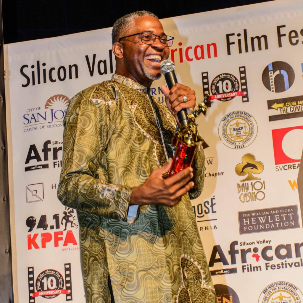 Un homme souriant en tenue traditionnelle tient un micro et un trophée devant une bannière du festival de film africain