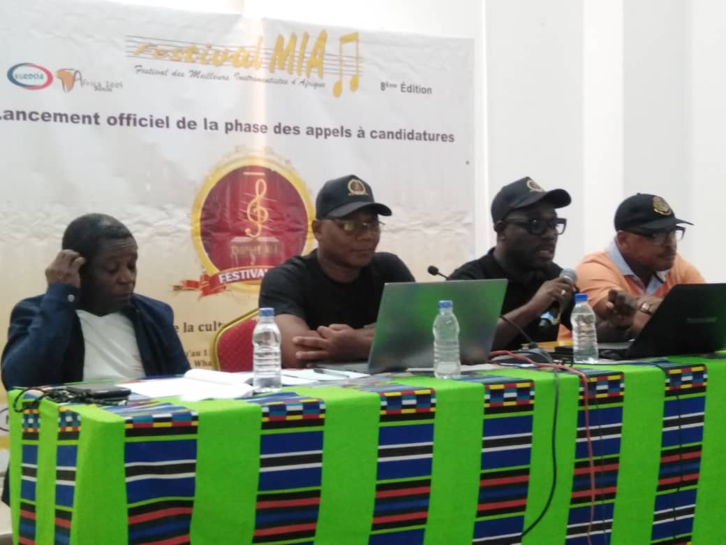 Quatre hommes assis derrière une table recouverte d’un tissu coloré avec microphones et ordinateurs lors d’une réunion