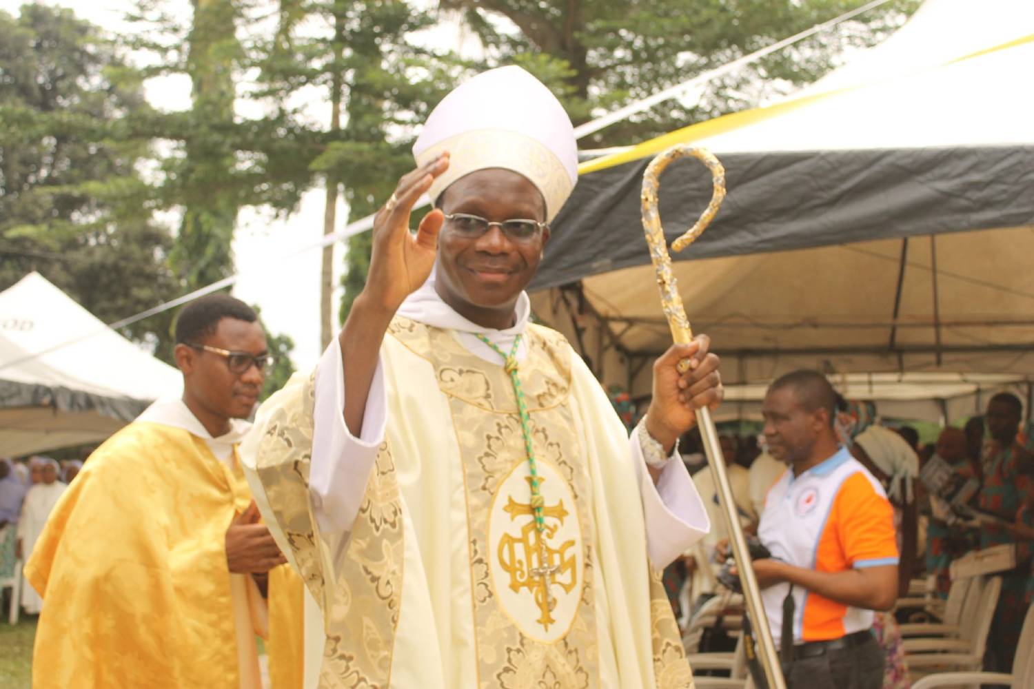 Un homme d'église en tenue liturgique blanche dorée, tenant une crosse, fait un geste de bénédiction en extérieur lors d'un événement.