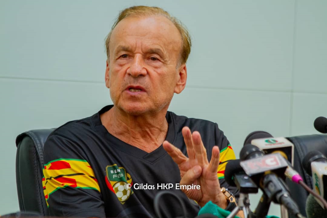 Homme discutant lors d une conférence de presse avec plusieurs micros devant lui et portant un maillot noir orné de bandes colorées sur les manches