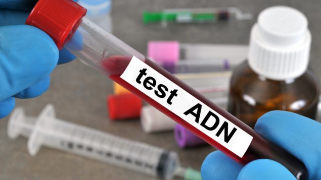 Une main gantée tient un tube rempli de sang étiqueté test ADN avec seringues et flacons en arrière-plan