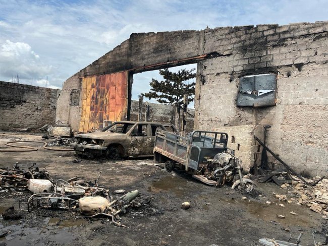Véhicules calcinés dans une cour entourée de murs en ruine sous un ciel partiellement nuageux