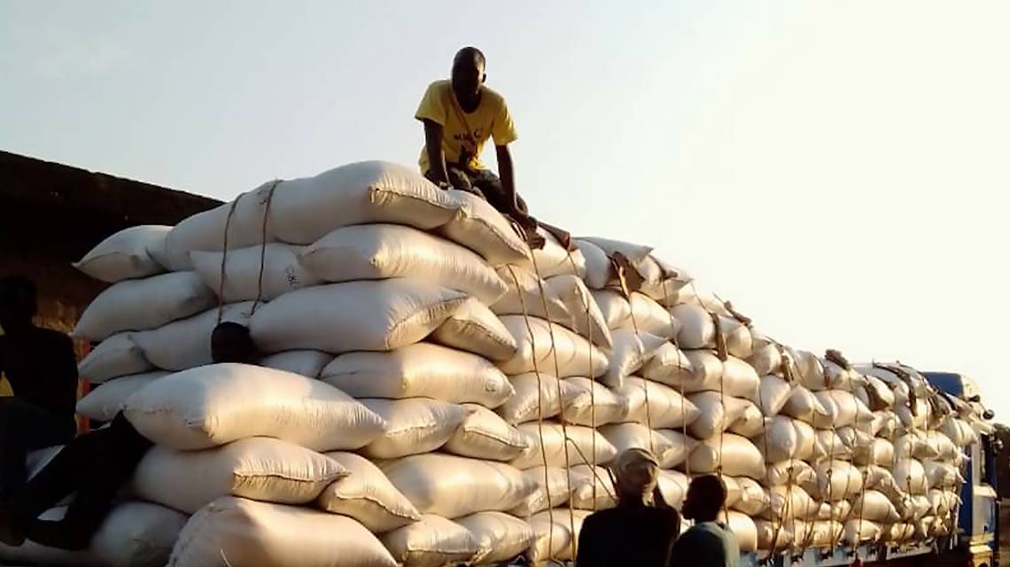 Un homme assis sur une grande pile de sacs blancs empilés sur un camion sous un ciel clair au coucher du soleil
