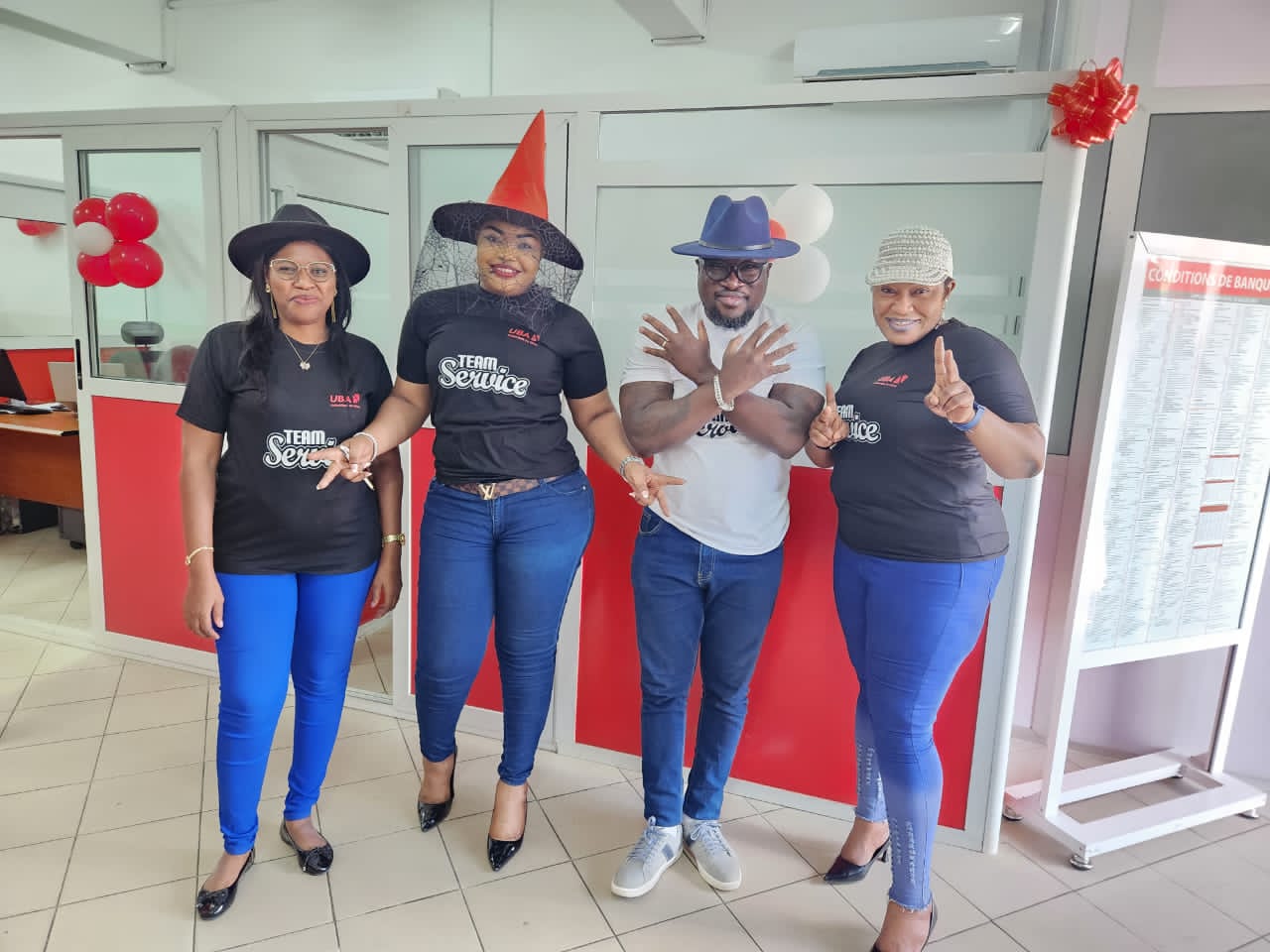 Quatre personnes debout en blouse noire et pantalon bleu, souriantes avec des chapeaux différents dans un bureau rouge et blanc