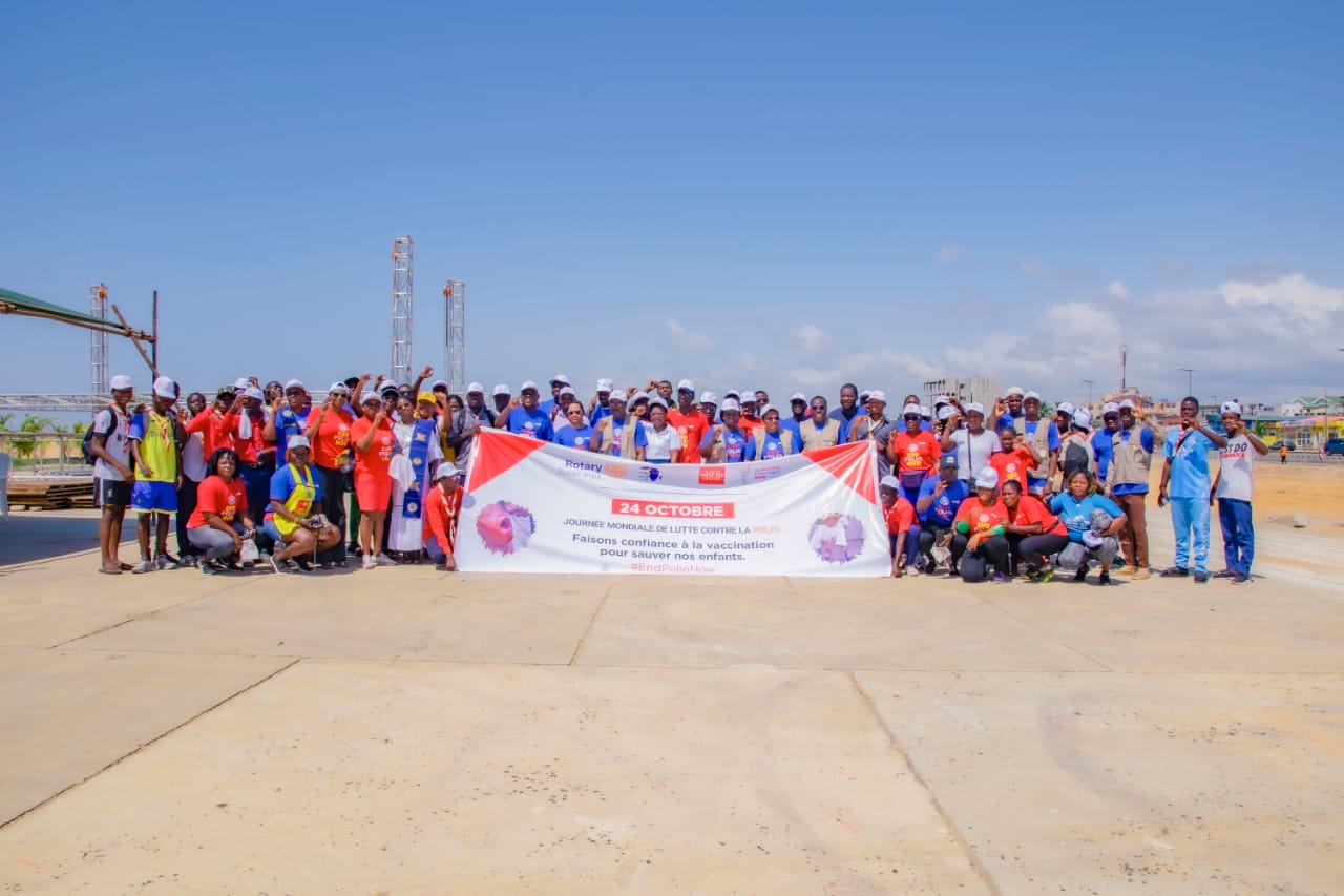 Groupe de personnes posant sur un terrain dégagé derrière une grande banderole blanche et rouge sous un ciel bleu avec quelques nuages.