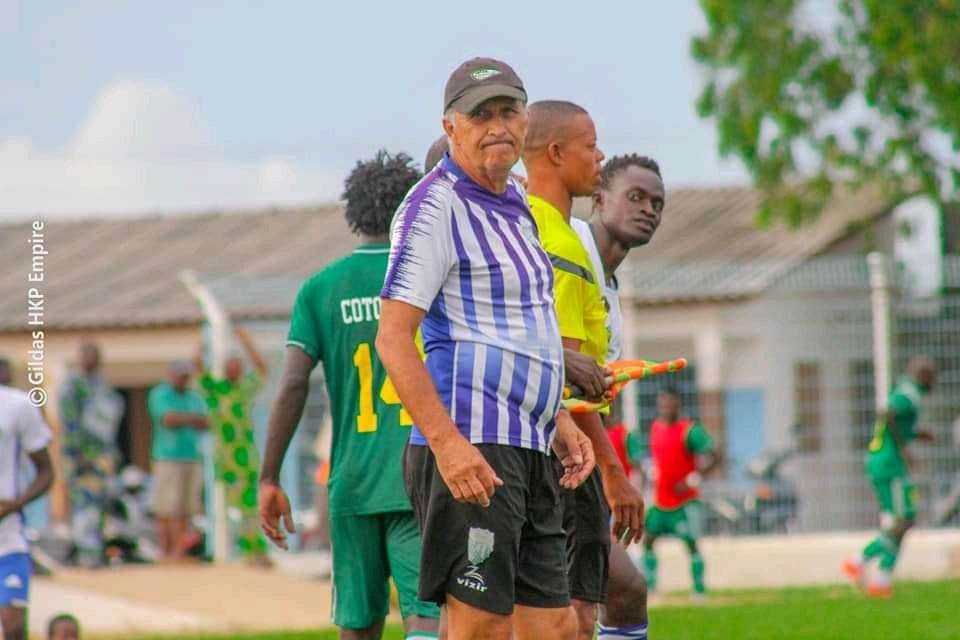 Un homme en tenue sportive rayée violette avec casquette noire sur un terrain de football avec joueurs en arrière-plan