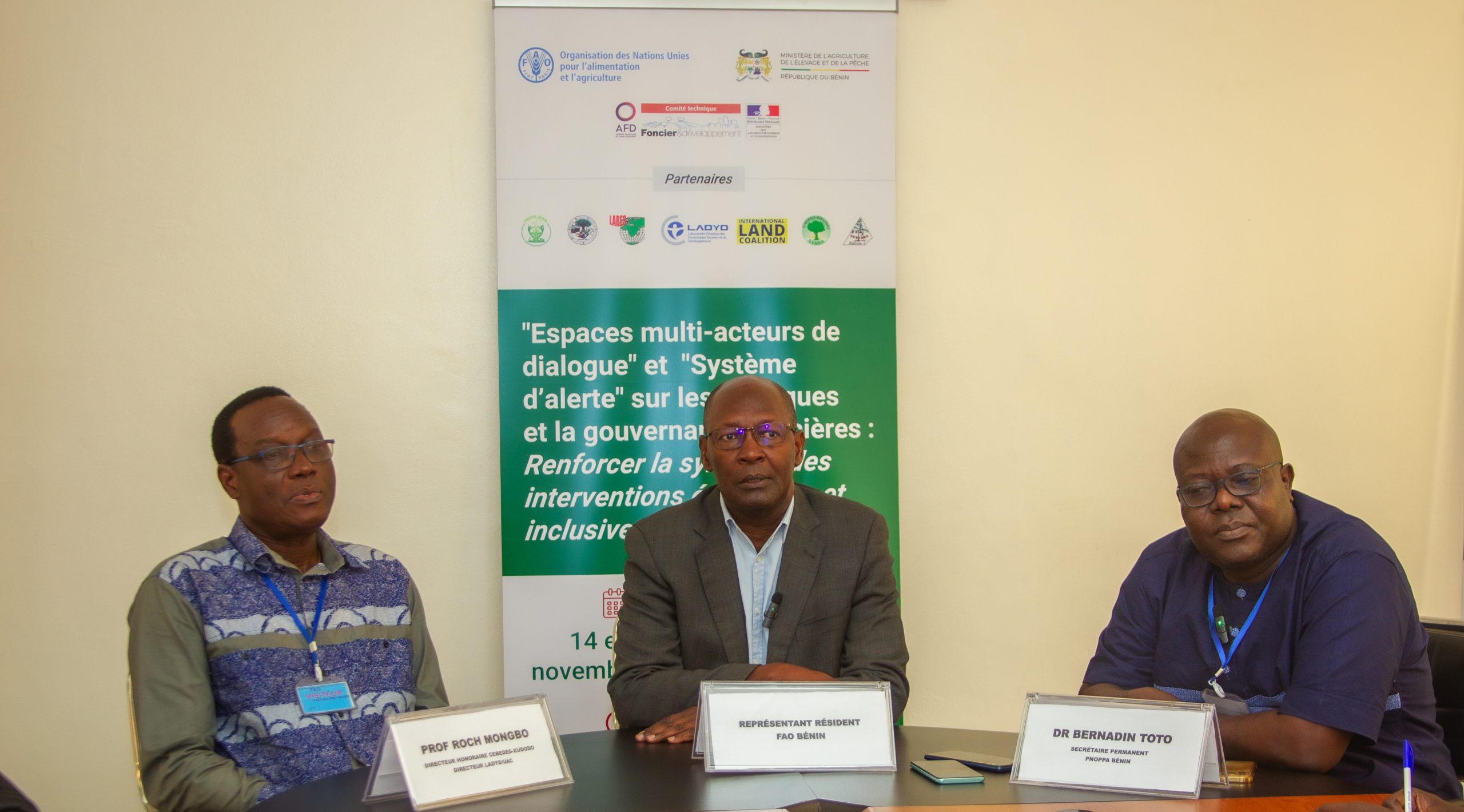 Trois hommes assis autour d'une table devant une affiche portant un texte sur la gouvernance foncière