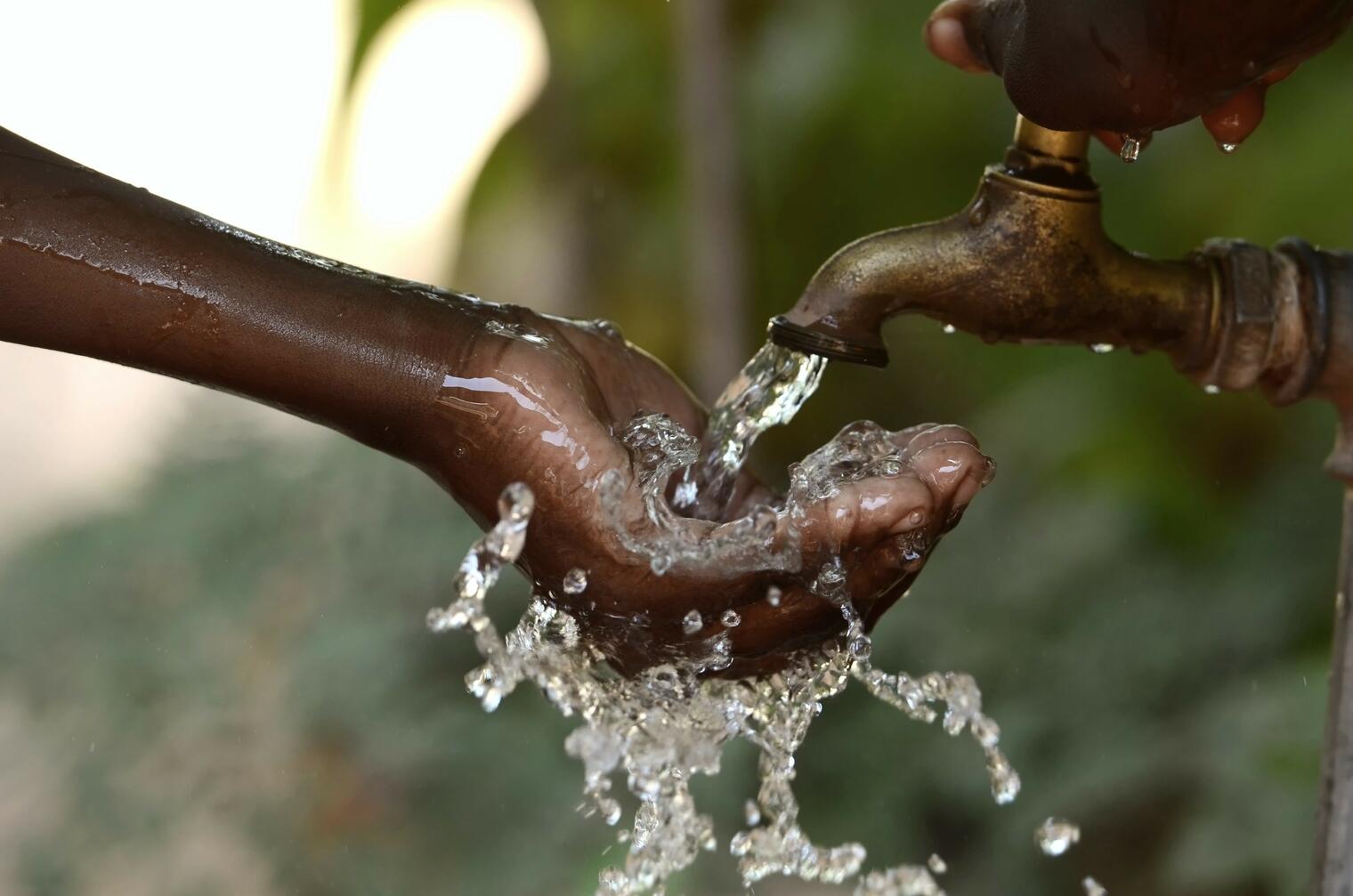 Une main reçoit de l'eau claire qui coule d'un robinet métallique dans un environnement extérieur flou
