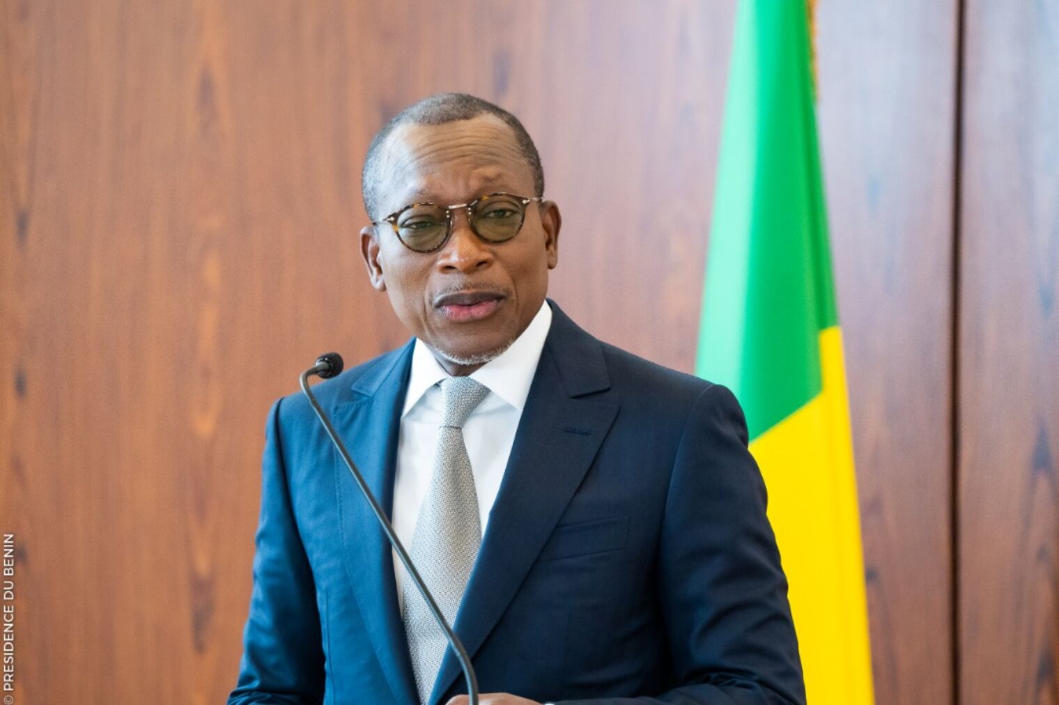 Homme en costume sombre et lunettes parlant au micro devant un drapeau vert jaune et rouge sur fond en bois