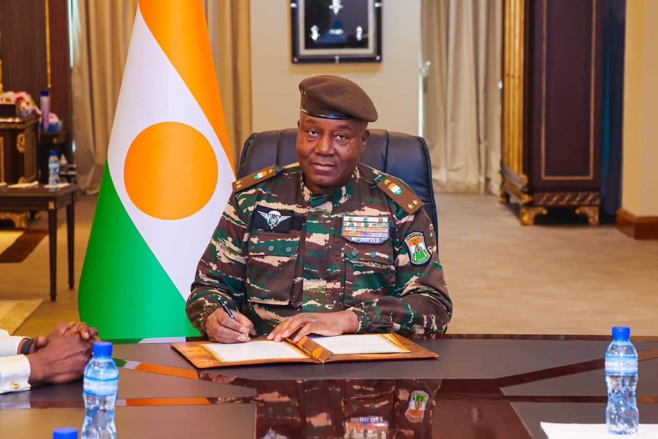 Un homme en uniforme militaire signe un document assis à une table avec un drapeau tricolore en arrière-plan
