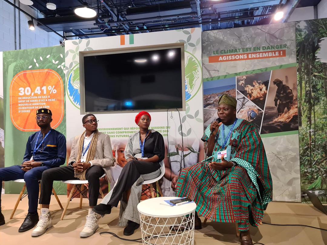 Quatre personnes assises en discussion avec une femme en tenue traditionnelle tenant un micro dans un espace d’exposition sur le climat