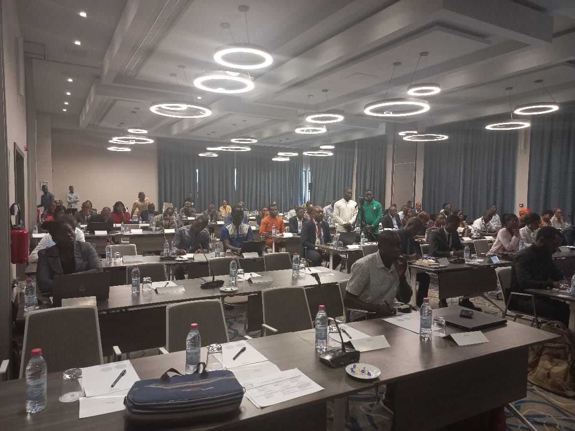 Salle de conférence avec des participants assis à des tables équipées de documents, bouteilles d'eau et micros sous des luminaires ronds modernes