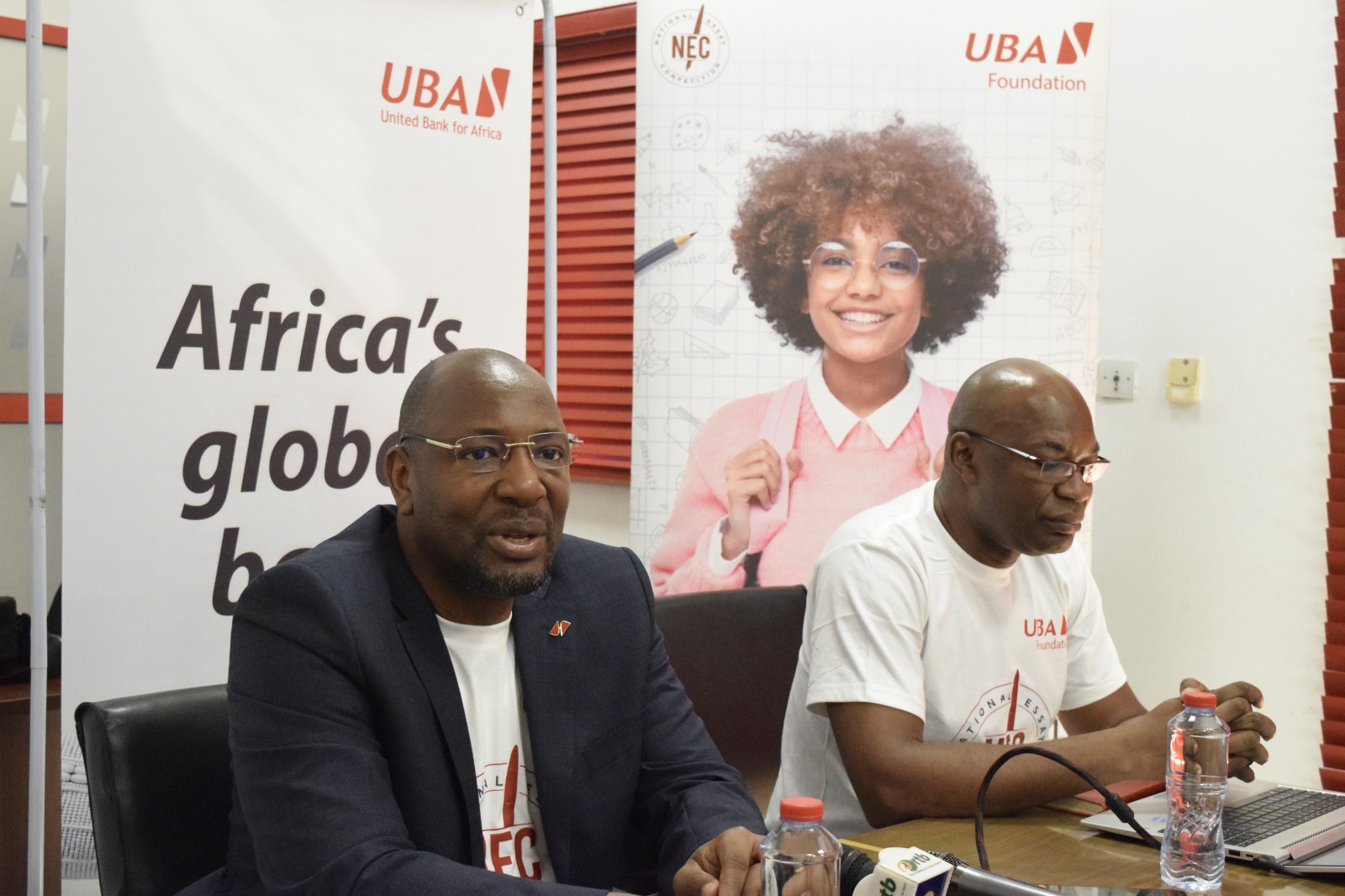 Deux hommes assis à une table avec microphones et bouteilles d'eau, devant des affiches promotionnelles d'une banque africaine