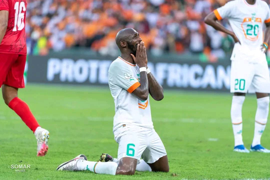 Joueur en maillot blanc agenouillé sur le gazon les mains sur le visage en signe de frustration pendant un match de football