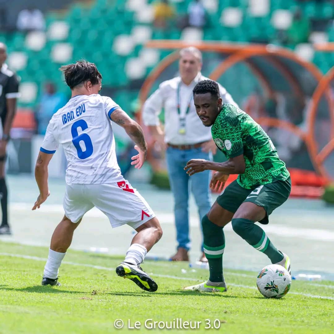 Deux joueurs de football en action, l'un en maillot vert contrôlant le ballon face à un adversaire en blanc sur un terrain gazonné