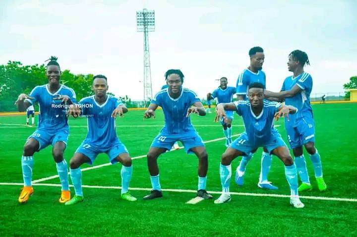 Sept joueurs en tenue de football bleue posent en souriant sur un terrain vert sous un ciel nuageux