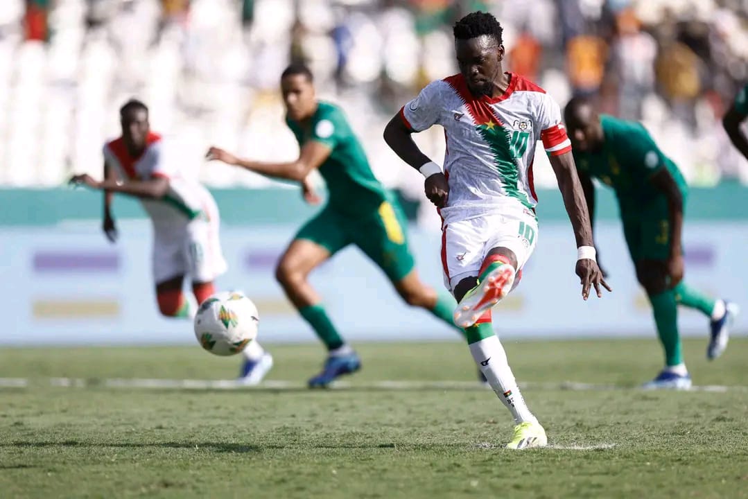 Un joueur en maillot blanc et rouge frappe le ballon devant des adversaires en vert sur un terrain de football.