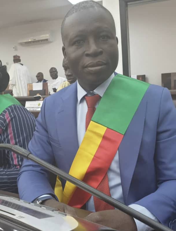 Un homme en costume bleu avec une écharpe tricolore rouge jaune vert assis dans une salle avec d’autres personnes