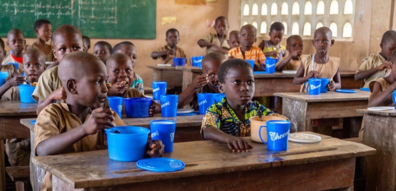 Des élèves d’une classe élémentaire sont assis à leurs bureaux en train de manger avec des bols et des gobelets bleus dans une salle de classe.
