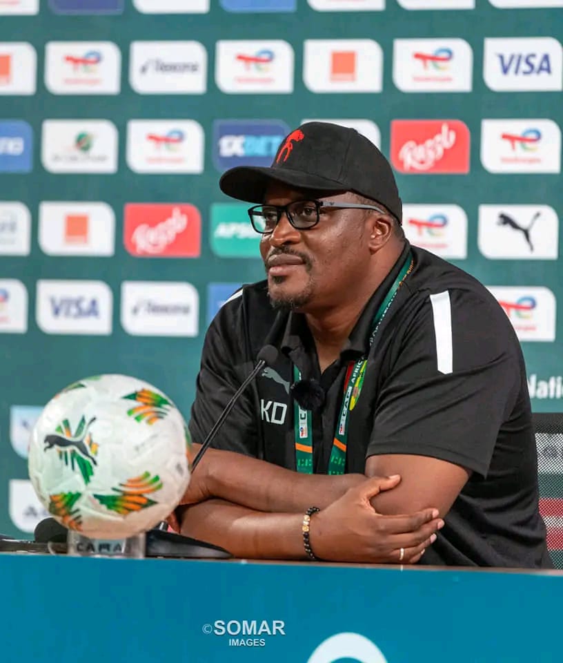 Un homme en polo noir et casquette noire avec lunettes parle lors d’une conférence avec un ballon de football sur la table devant lui