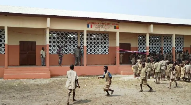 Enfants en uniforme jouant devant un bâtiment scolaire à l'extérieur avec plusieurs adultes présents à l'entrée