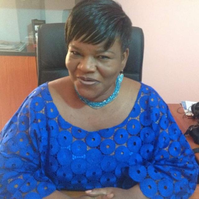 Femme souriante en tenue traditionnelle bleue assise dans un bureau devant un meuble et un ordinateur.