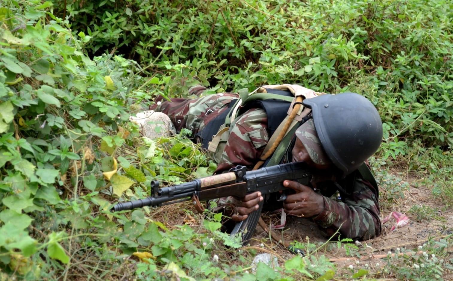 Un soldat en uniforme et casque noir est allongé dans la végétation avec un fusil d'assaut en position de tir