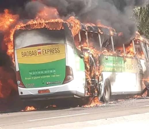 Un bus est entièrement en flammes, avec de la fumée noire s'échappant du toit et des côtés sur une route.